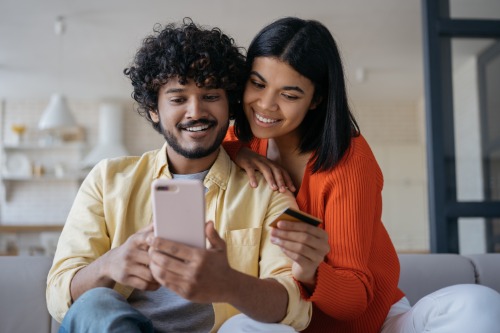 man and woman with dark black hair sitting on a couch looking at his phone. She has a credit card in her hand. 